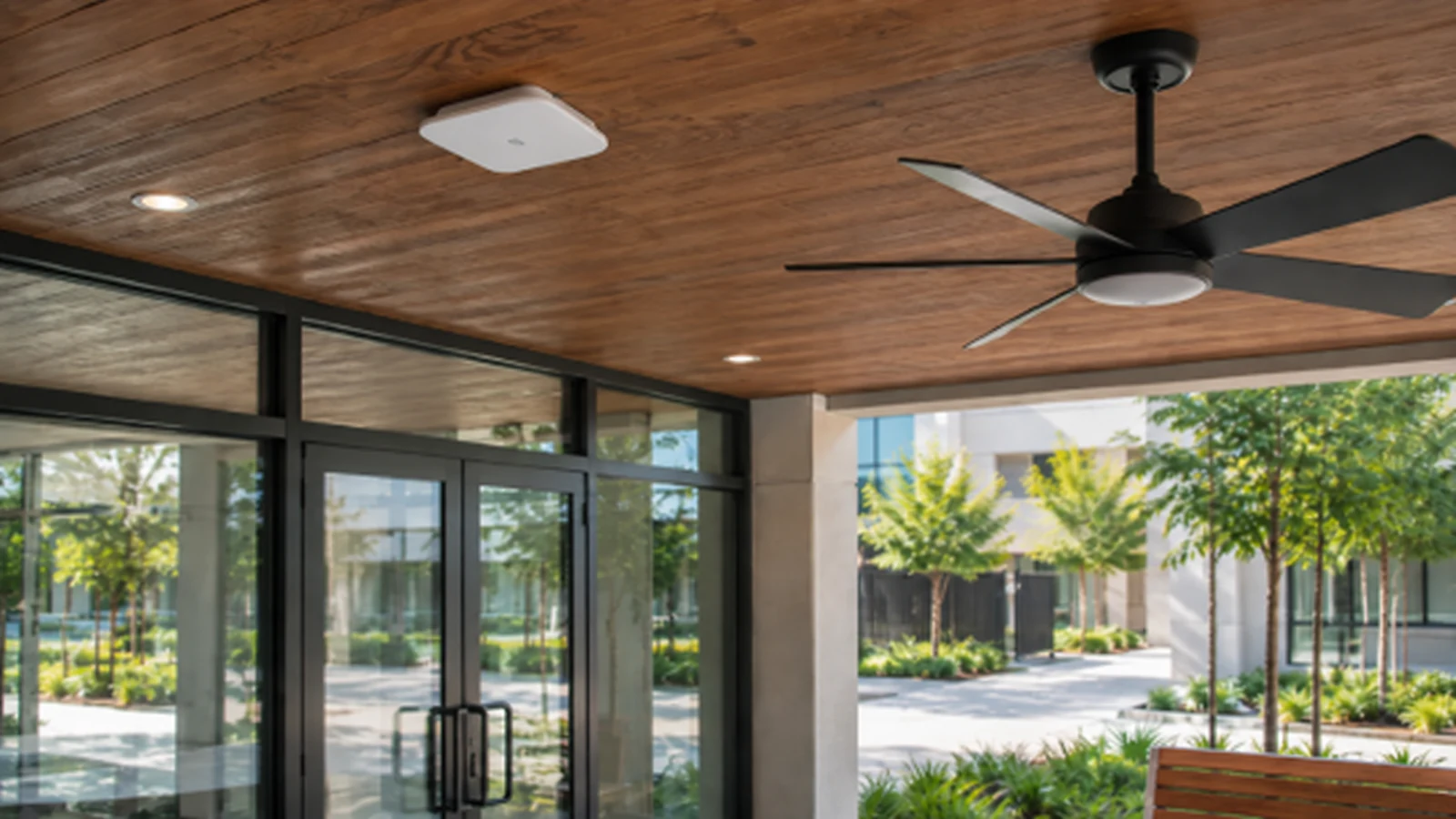 Commercial wireless access point mounted on a wood soffit beside a ceiling fan and glass entrance